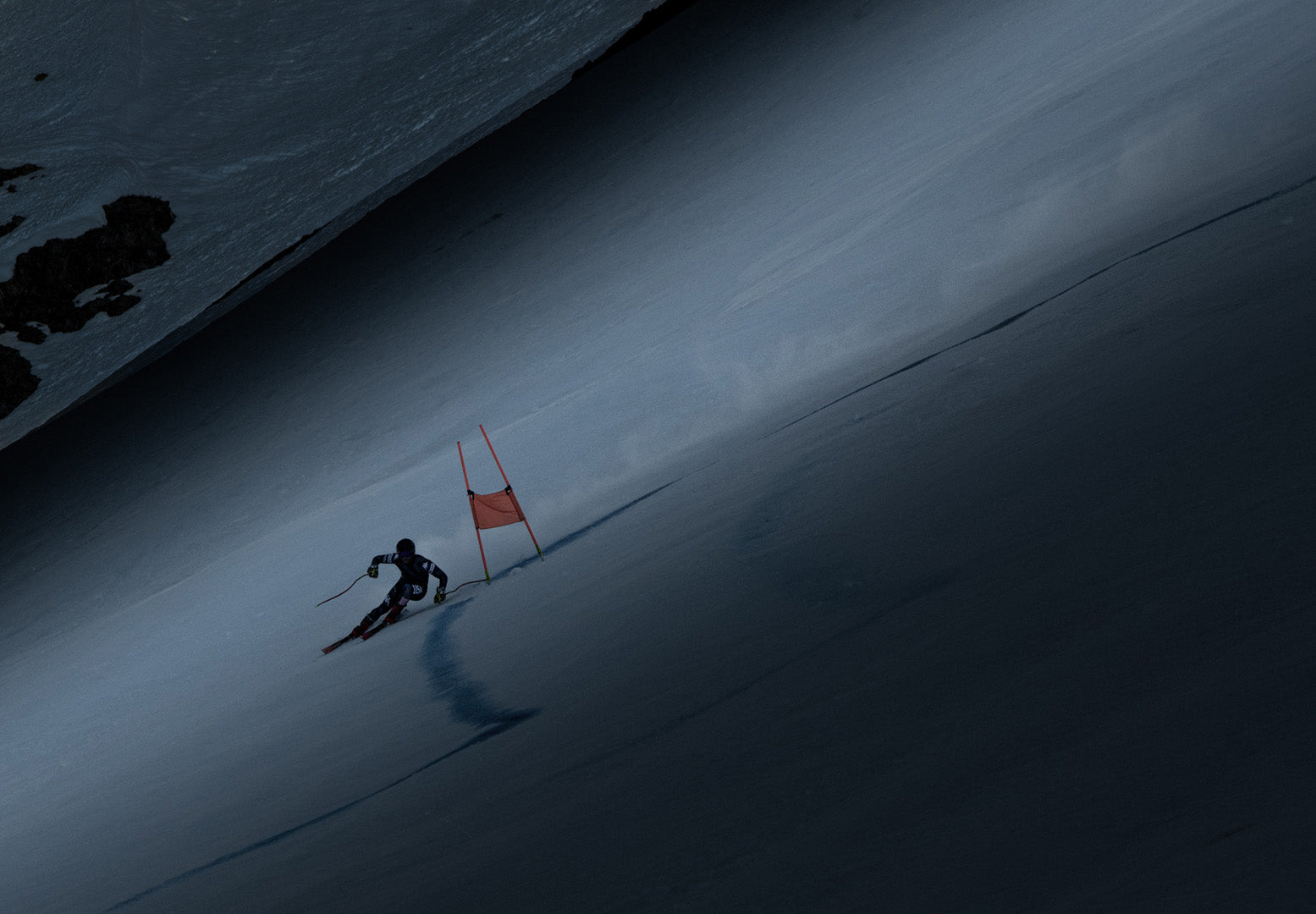 Darkly lit dramatic image of US ski athlete carving across steep mountain next to an orange ski gate.