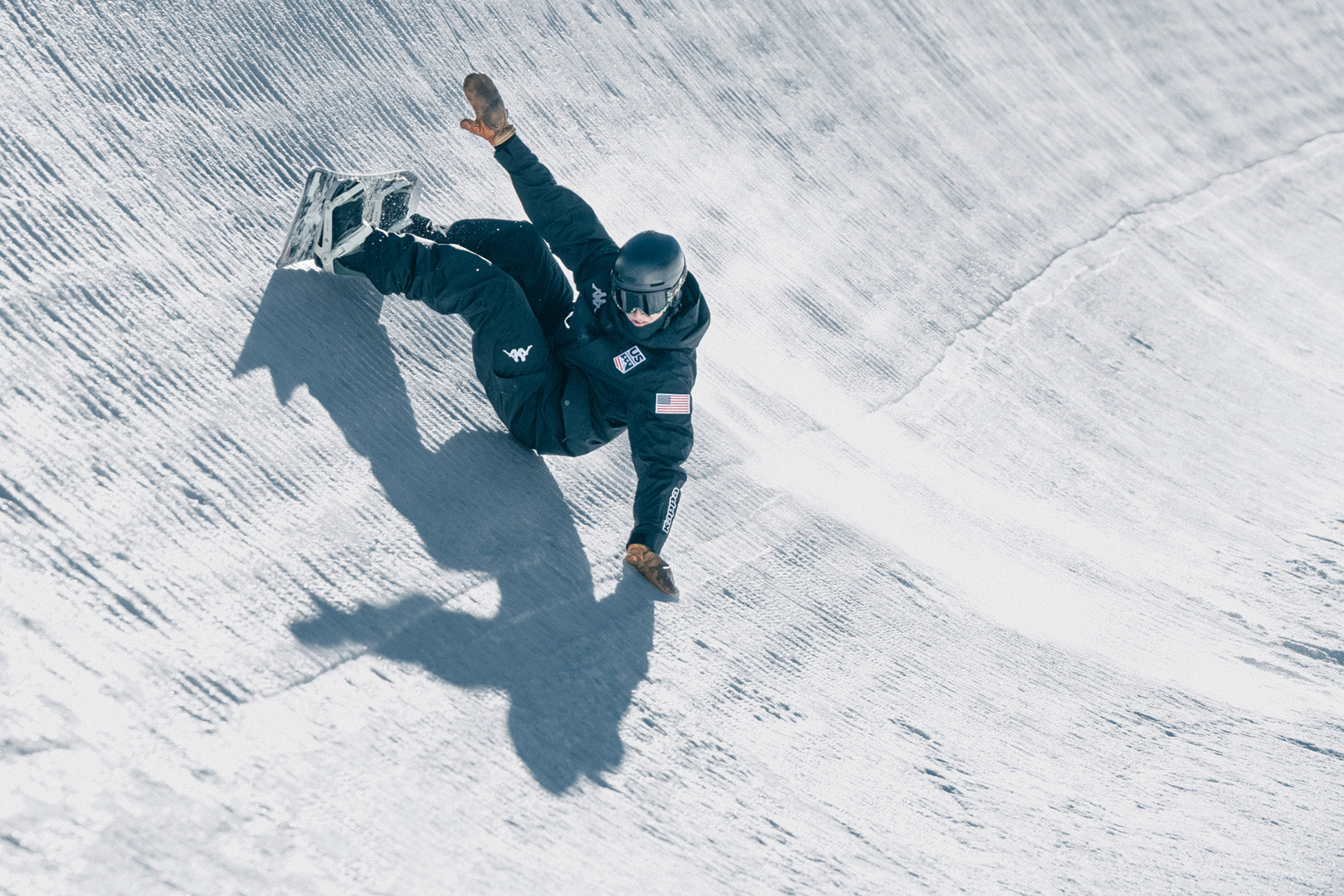 Male US snowboard athlete carving half pipe with his hand touching the snow in full Kappa uniform.