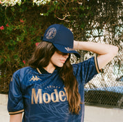 Female model wearing navy blue cap with Modelo logos in gold on bill and side of cap.