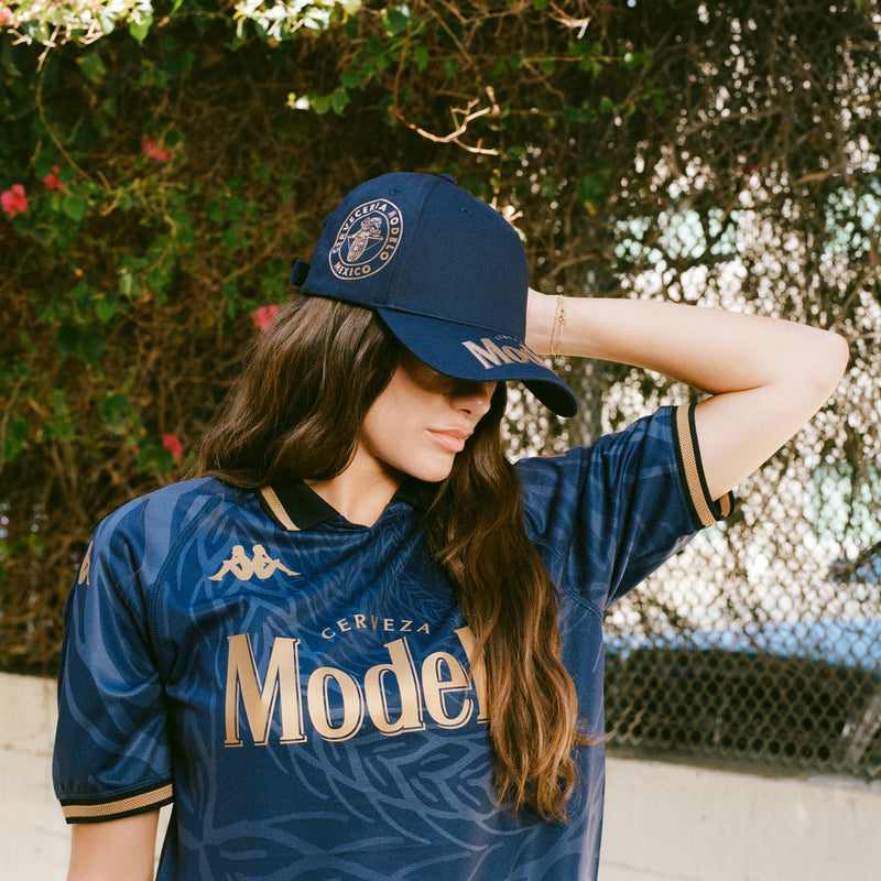 Female model wearing navy blue cap with Modelo logos in gold on bill and side of cap.