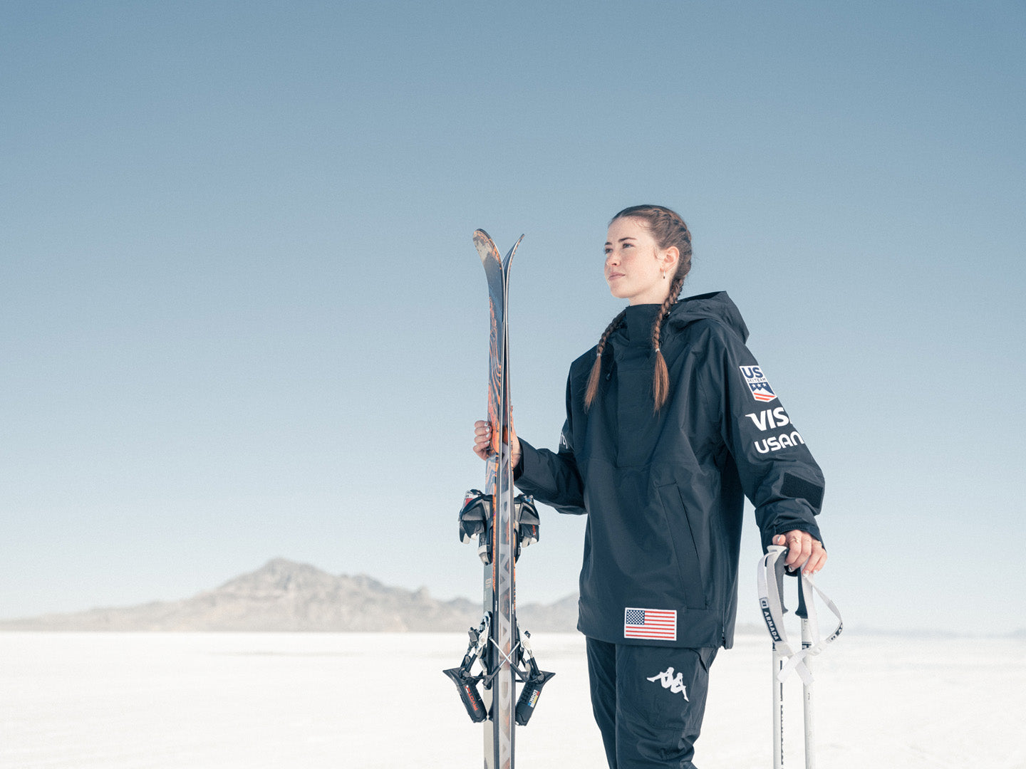 Female US ski athlete holding ski in her right hand, poles in her left hand, standing on salt flats looking off into the distance