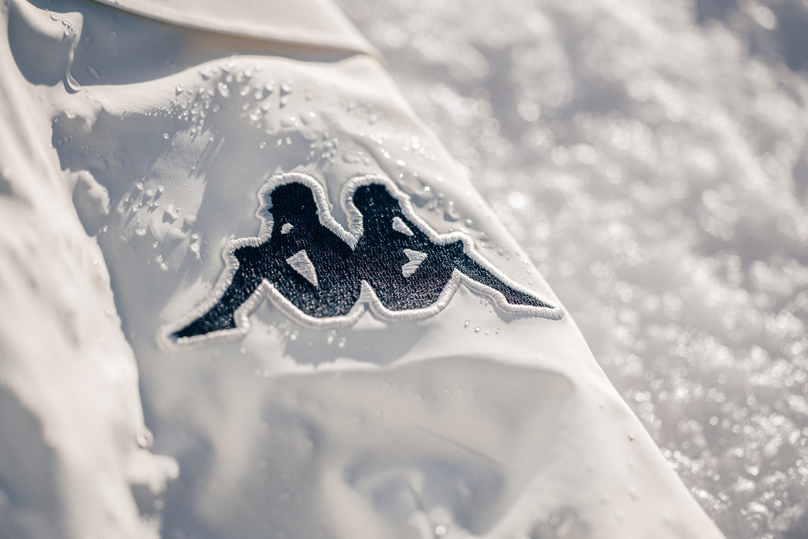 Extreme close up of white Kappa ski jacket with dew and powdery ice surface. Embroidered Logo in focus.
