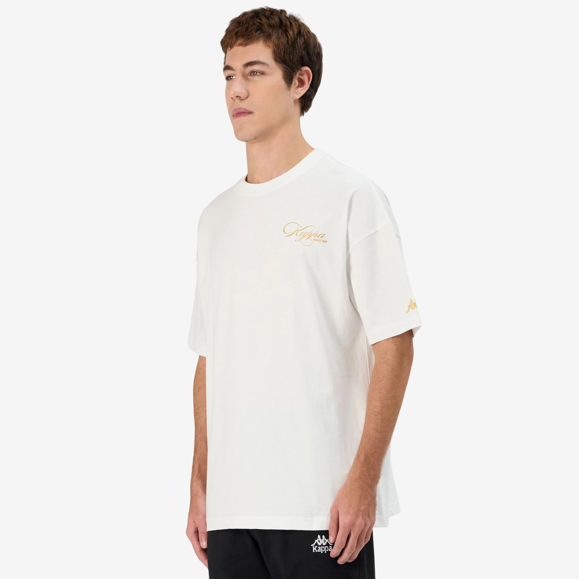 Man wearing white Kappa t-shirt with gold script on front left side and gold omini logo on left sleeve. Side view.